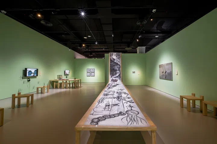 展览现场照片：新加坡美术馆《The Living Room》，中央是一条长桌，上面铺满黑白绘画卷轴，周围墙面展示多件影像与绘画作品，空间以浅绿色墙面营造沉静氛围。在新加坡去哪里玩？新加坡好玩推荐