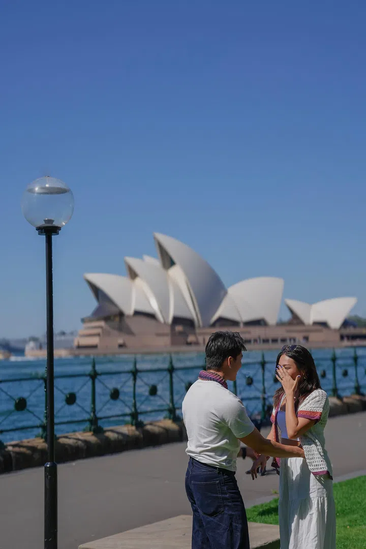 在澳洲悉尼歌剧院前的户外海滨步道上，Edwin Goh与Rachel Wan面对面站立，Rachel以手掩面露出惊喜神情，画面洋溢求婚瞬间的感动氛围，背景是蓝天与标志性的悉尼歌剧院建筑。Rachel Wan：最好的感情，是让我更像自己