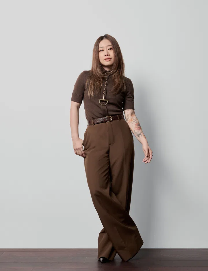 新加坡植物壁画艺术家 Geraldine Toh 身穿深棕色针织上衣与阔腿裤，站在浅灰色背景前。她以自然为创作灵感，通过壁画为城市空间注入绿意与宁静。