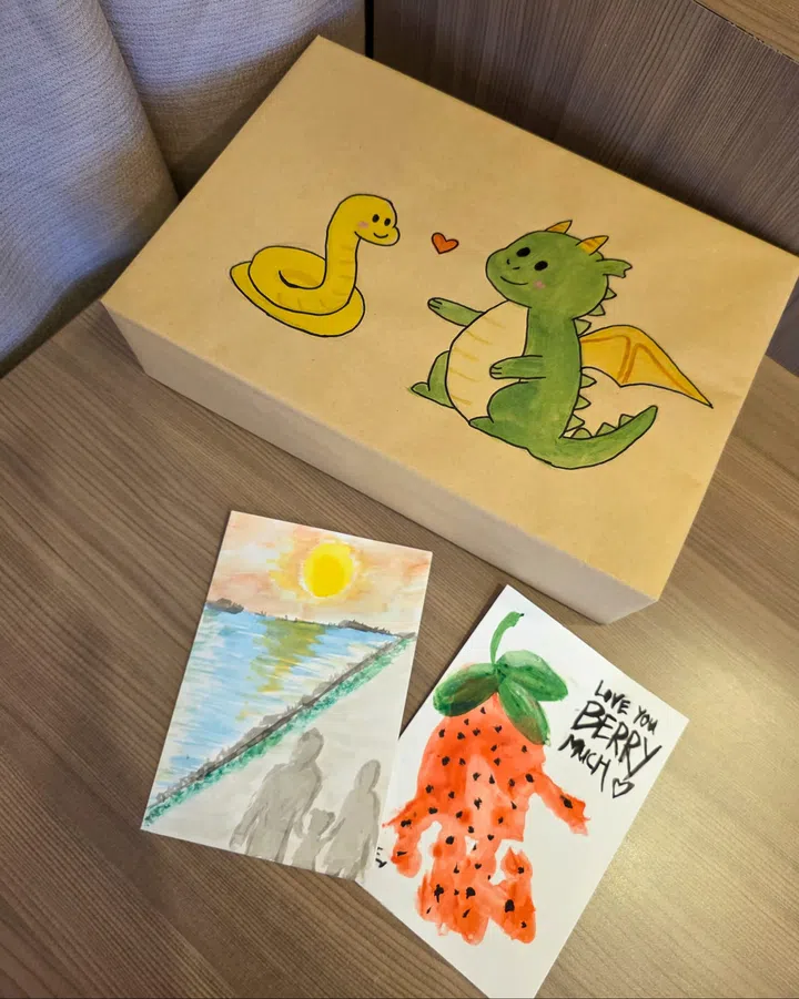 画面中有一只手绘礼盒，上面画着可爱的绿色龙与黄色蛇，中间有红色爱心；桌上摆着两幅水彩画，一幅是日落下的亲子剪影，一幅是草莓图并写着“Love you berry much ♥”。作品展现陈泂江一家以艺术表达亲情的温馨日常。陈泂江独家专访。