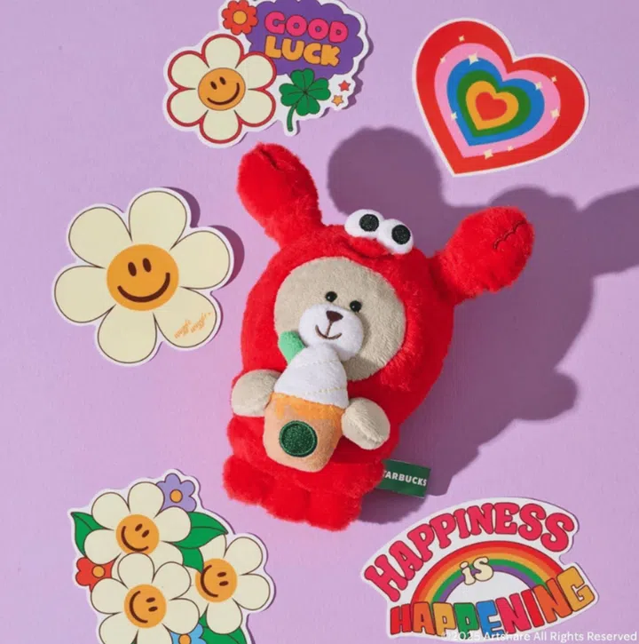 Starbucks 与 Wiggle Wiggle 联名系列周边展示图，画面中央为 Starbucks Bearista 公仔，身穿 Wiggle Wiggle 经典红色龙虾造型并手持咖啡杯，背景搭配彩色花朵、彩虹爱心与 ‘Happiness is Happening’ 图案贴纸，整体呈现充满童趣与色彩感的限量联名公仔周边，适合喜爱角色联名与设计感生活小物的读者。