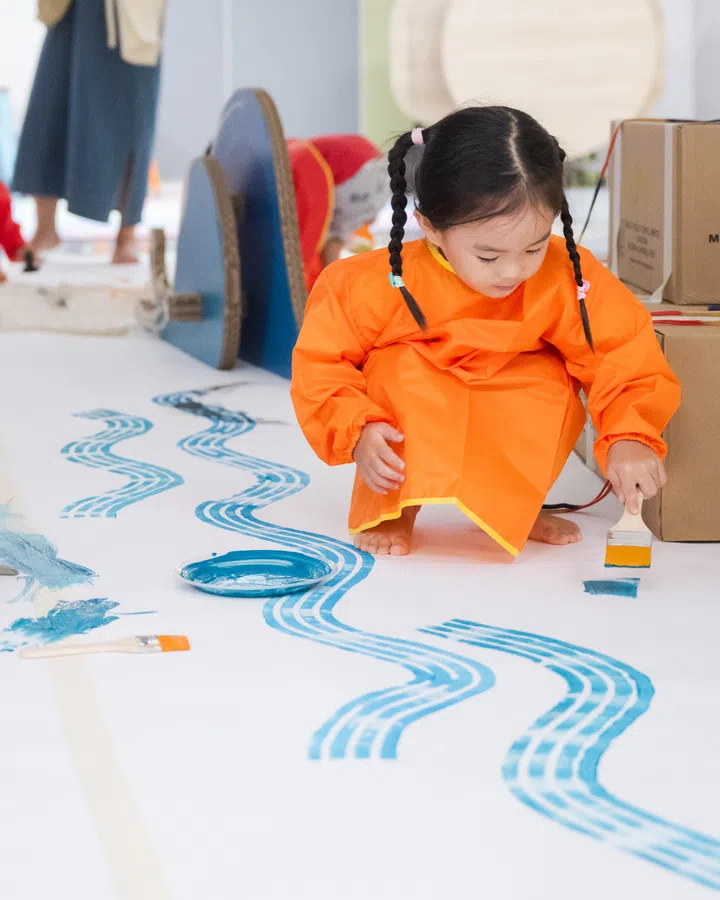 一名穿着橙色画衣的小女孩在地上作画，用蓝色颜料和画笔创作波浪图案，展现 National Gallery Singapore 十周年庆的亲子艺术活动氛围。在新加坡去哪里玩？新加坡好玩推荐