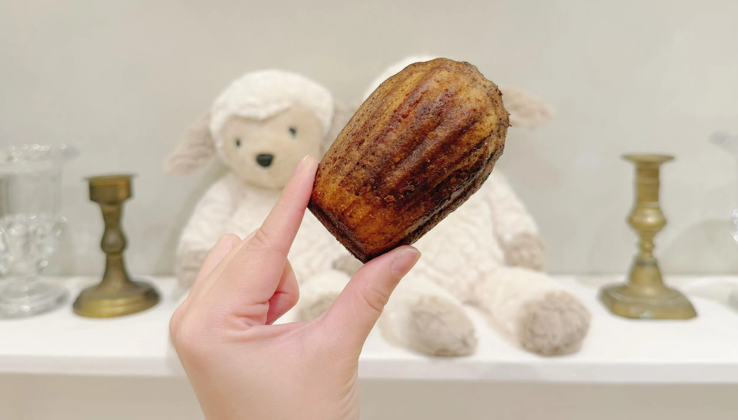 手持一块外层金黄焦脆、带有烘烤纹路的韩国脆皮牛油年糕（Butter tteok），近距离展示其外脆内软的质感，背景为温馨居家摆设与毛绒玩偶，呈现近期爆红K-food甜点的视觉特色。