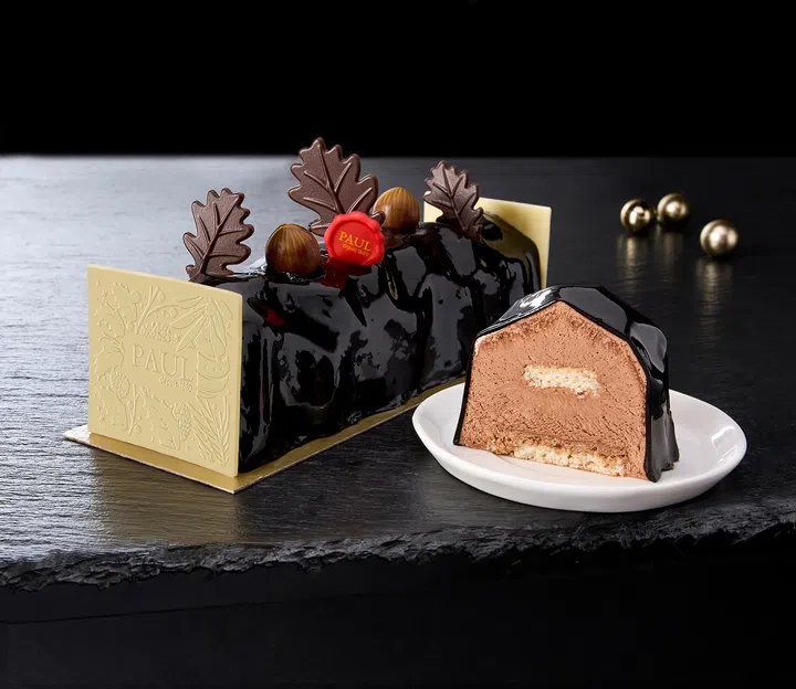 PAUL 的 Midnight Chocolate Noël 圣诞蛋糕，镜面黑巧克力外层搭配巧克力叶片与红色装饰，切片展示绵密巧克力慕斯与饼层结构，整体摆放在深色石板背景中。2025 新加坡圣诞蛋糕推荐｜好吃又好看的 Christmas Cakes & Logcakes 精选清单