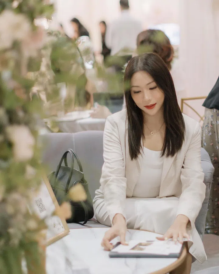 Rachel Wan身穿白色上衣与浅色西装外套，坐在室内优雅空间里翻阅相册或资料，桌旁放着深绿色手袋与花艺装饰，神情专注温柔，整体氛围宁静而精致。Rachel Wan：最好的感情，是让我更像自己