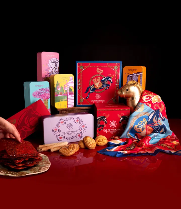 画面展示 Old Seng Choong 马年新春系列年饼与礼盒陈列在红色桌面上，背景为深色渐层，整体营造浓厚节庆氛围。中央与后方摆放多款不同颜色与图案设计的铁盒与礼盒包装，包括红色马年主题礼盒、粉色与绿色插画风格铁盒，以及印有传统建筑与自然元素的黄色铁盒。前景左侧是一盘叠放整齐的肉干，有一只手正从盘中取起一片肉干，呈现拜年招待亲友的情境。桌面中央摆放多款传统年饼点心，包括鸡蛋卷与中式酥饼。右侧可见一只金色马造型摆饰，旁边铺放着蓝红色新春图案丝巾，呼应马年主题与送礼搭配元素。整体画面呈现马年新春年饼从肉干到传统点心与节庆礼盒的多样选择，适合拜年与新春送礼场合。文章标题：“2026马年年饼推荐：新春送礼与拜年点心精选”。