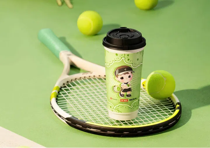 一杯印有Hacipupu网球小冠军造型的CHAGEE青提奶茶放在网球拍上，周围散落着网球，整体以绿色为主调，清新活泼，充满运动与可爱氛围。