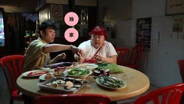 电影《A Good Fortune》《百万红包》中的一幕，两人坐在老式小贩中心风格的小店圆桌旁，桌上摆满切好的茄子、秋葵、辣椒、青菜与豆腐等家常食材，正一起准备料理。红色塑料椅与墙上的饮料与禁烟标志营造出本地小店的生活氛围，画面呈现出温馨、真实的日常用餐时刻，呼应电影以家庭与快乐为主题的故事基调。黄伟良林茜茜 WANG WEI LIANG AND XIXI LIM