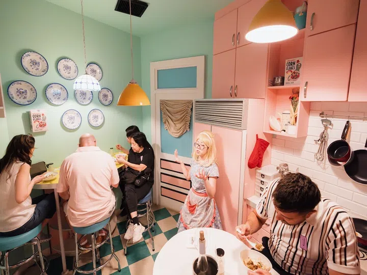 几位参与者坐在复古风格的厨房中用餐，墙上装饰着蓝白瓷盘与粉红橱柜，一位金发演员在一旁表演互动，为《Absurdities》沉浸式用餐体验增添戏剧氛围。哪里可以用SG Culture Pass？文化通行证活动推荐