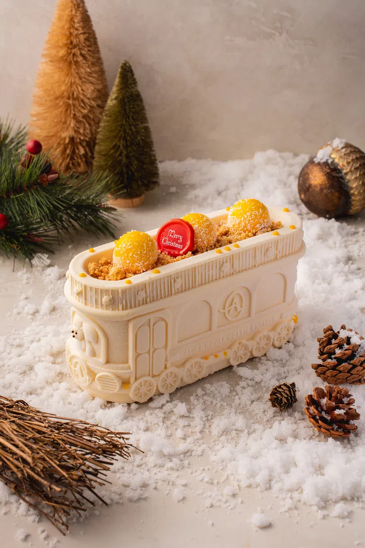 Alice Boulangerie 的圣诞火车造型蛋糕，雪白外层搭配黄色果球与红色“Merry Christmas”装饰，置于松果与人造雪景中，呈现浓厚节日氛围。2025 新加坡圣诞蛋糕推荐｜好吃又好看的 Christmas Cakes & Logcakes 精选清单