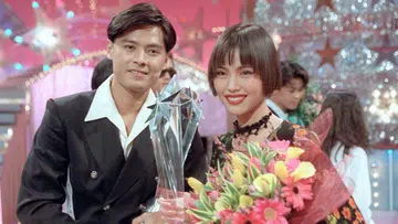 颁奖典礼现场，一名男艺人身穿黑色西装手持奖座，与一名短发女艺人并肩合影，女艺人手捧鲜花，两人面带笑容，背景为灯光闪耀的舞台装饰。李南星与郑惠玉