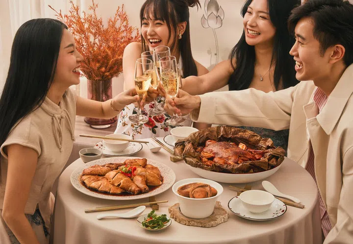 农历新年团圆用餐场景展示图，四位年轻人围坐在圆桌旁举杯庆祝，桌上摆放多道节庆佳肴，包括以荷叶包裹的八宝鸭、切片酱油鸡、鲍鱼汤盅及多款配菜，整体气氛温馨热闹，呈现朋友或家人共享团圆饭的新加坡农历新年外卖与节庆聚餐氛围。