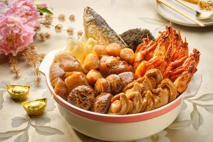 白色砂锅中盛装新春盆菜，整齐铺排整虾、鲍鱼、干贝、花胶、香菇、发菜与一整条鱼，食材表面淋上浓亮酱汁，旁边点缀金元宝摆饰、花卉与中式餐具，营造喜庆隆重的农历新年团圆饭氛围，新加坡盆菜推荐2026 农历新年必吃餐厅与价格整理。