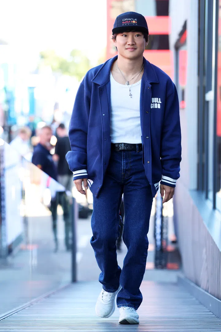 照片中，角田裕毅（Yuki Tsunoda）头戴 Oracle Red Bull Racing 黑色帽子，身穿蓝色外套、白色背心和牛仔裤，脚踩白色运动鞋，走在赛场通道上，神情轻松。他是F1年轻的亚洲车手之一。F1赛车手Yuki Tsunoda角田裕毅参加2025新加坡F1 Grand Prix