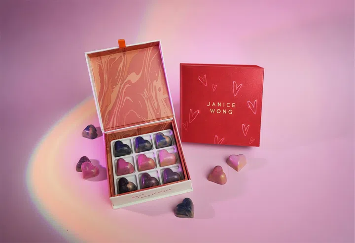 Janice Wong 情人节限定巧克力礼盒摆放在粉色背景上，整体画面呈现梦幻而精致的节日氛围。礼盒打开后，可见内部整齐排列九颗心形巧克力，每颗巧克力呈现渐变色泽，包括深紫、玫粉与金属光泽的色调，外观如艺术品般精致。礼盒内侧为粉色大理石纹设计，增添视觉层次与艺术感。礼盒旁摆放红色包装盒，上面印有“JANICE WONG”字样与手绘爱心图案，强化情人节主题。周围散落几颗心形巧克力，使画面更具动感与立体感。柔和光线在背景形成淡淡彩虹光晕，营造出浪漫且现代的氛围，突显礼盒作为情人节巧克力礼物的独特设计与精致质感。2026情人节礼物推荐：10款精致礼物让TA一秒心动