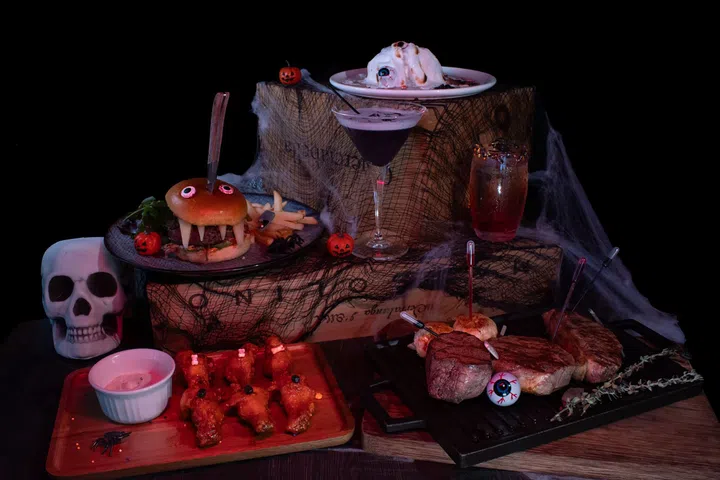 摆放在桌上的万圣节主题料理，包括插着刀的怪兽汉堡、火红鸡翅、牛排与暗黑鸡尾酒，营造诡异又趣味的节日氛围。新加坡万圣节去哪里玩推荐。