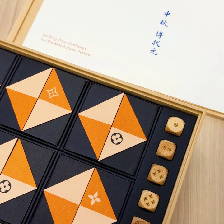 Louis Vuitton“中秋博状元”礼盒局部特写，浅木色外框内排列深蓝色方盒，盒面以橙色与浅木色三角形拼成菱形，并点缀Monogram花卉图案。右侧凹槽整齐放置木色骰子，骰面以品牌花卉纹样组成不同点数。上方白色说明页印有蓝色“中秋博状元”题字及英文游戏名称。