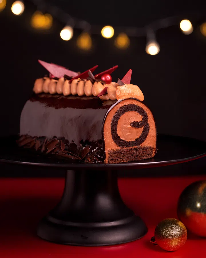 Cat & the Fiddle 的 Dark Chocolate Log Cheesecake，表层覆上亮泽黑巧克力，中间以巧克力螺旋切面呈现浓郁质地，顶部点缀红色与巧克力装饰，置于黑色蛋糕架与圣诞摆设中。2025 新加坡圣诞蛋糕推荐｜好吃又好看的 Christmas Cakes & Logcakes 精选清单