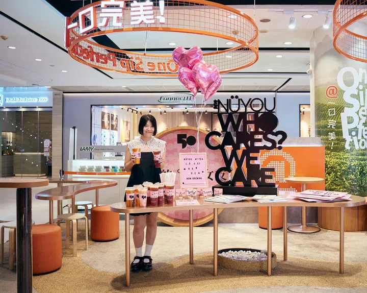 《女友》总编辑艳玲站在“NÜYOU WHO CARES? WE CARE.”主题装置前，双手分别拿着两杯“女友莓果伯爵”联名茶饮。前方桌面摆放多杯联名饮品、《女友》杂志及活动展示物，背景可见粉色拍照装置、粉色爱心气球及《女友》50周年主题布置，以粉色和橙色为主色调。