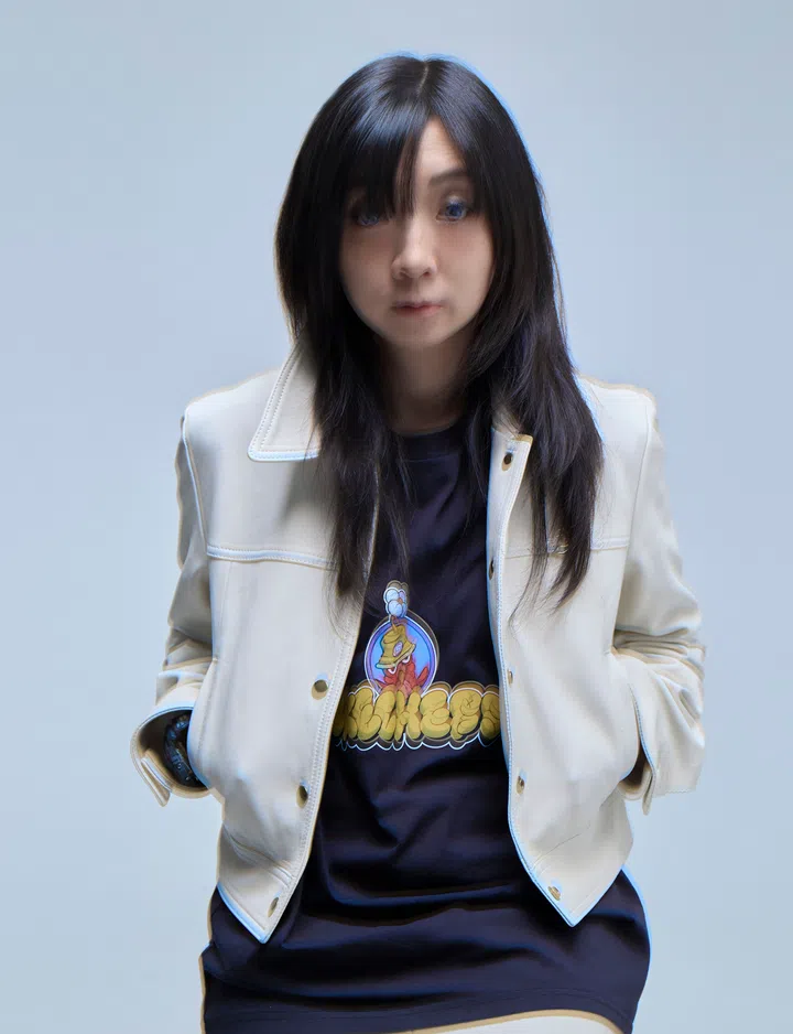 新加坡插画师Erika Tay身穿Skechers联名系列黑色T恤，外搭COACH的浅米色皮质外套，双手插袋，自信站立。T恤正面印有Skechers联名卡通Logo图案，整体造型简约又带有街头潮流感，展现Erika Tay的个人风格与联名系列的时尚氛围。