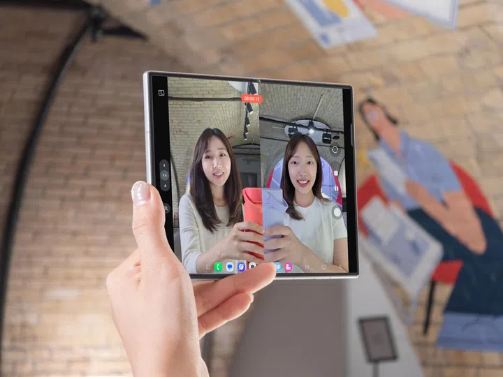 一只手握着展开状态的Samsung Galaxy Z Fold8，正在使用后置镜头进行自拍录影。折叠屏幕采用分屏预览模式，左侧显示摄影师透过后置镜头拍摄的画面，右侧则同步显示被拍摄者的即时预览，让双方都能同时确认构图与拍摄效果。画面中两位年轻女性分别手持粉红色及淡紫色水壶，对着镜头微笑，背景为室内砖墙空间。手机屏幕上方可见录影计时画面，整体展示Galaxy Z Fold8利用折叠屏幕实现双预览（Dual Preview）及后置镜头自拍录影功能，提升多人拍摄与内容创作时的便利性。