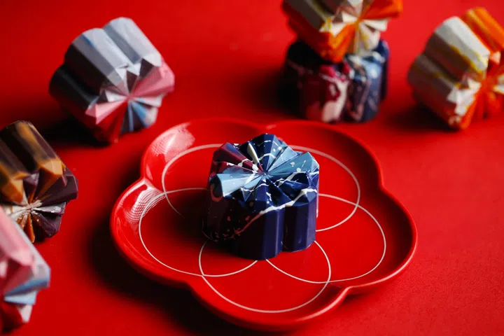 一枚蓝色几何造型的 Janice Wong 巧克力月饼置于红色花瓣形餐盘上，表面带有泼墨般的白色与浅蓝色纹理。周围散落着不同颜色的月饼，包括橙色、紫色与粉色，整体以红色背景衬托，呈现浓烈的视觉冲击感。Janice Wong, 2025中秋月饼必买推荐