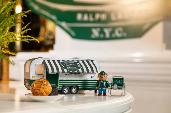 Ralph’s Coffee 2026中秋月饼礼盒场景图，画面中央为以品牌招牌绿白配色打造的复古露营车造型礼盒，车身印有Ralph’s Coffee与Ralph Lauren标志。礼盒旁摆放身穿绿色制服的Polo Bear-rista公仔，以及迷你露营椅与咖啡杯装饰；前景白色小碟上陈列一颗金黄色月饼与一块深色甜点。背景可见Ralph’s Coffee咖啡店的绿色遮阳篷，整体营造轻松、复古又具收藏感的中秋氛围。