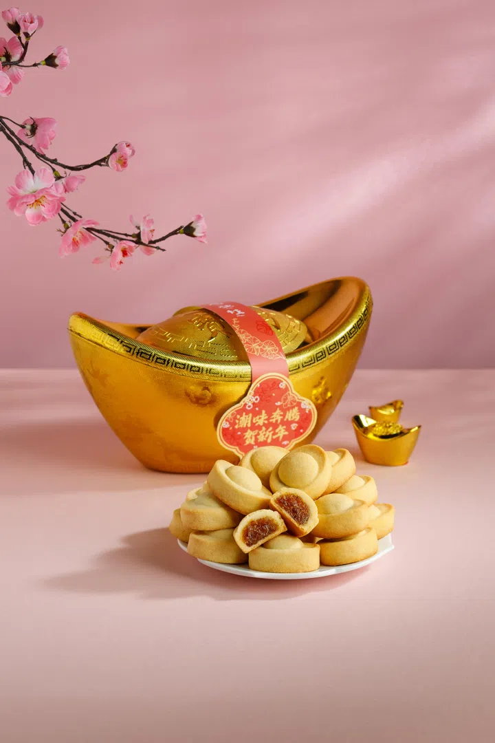 画面以粉色背景呈现新春吉祥意象，前景白色小盘上整齐堆叠多枚元宝造型凤梨酥，其中一枚被切开，露出金黄色凤梨内馅，突出酥皮与果馅的层次口感。后方摆放大型金色元宝装饰摆件，元宝上系有红色新春祝福吊牌，写有贺年祝语，象征招财进宝与新年好运。右侧另有迷你金元宝摆饰点缀，左上角可见粉色梅花枝垂入画面，为整体构图增添节庆与春意氛围。整体画面以传统吉祥元素搭配年饼点心，呈现适合新春拜年与送礼的节庆甜点意象。文章标题：“2026马年年饼推荐：新春送礼与拜年点心精选”。