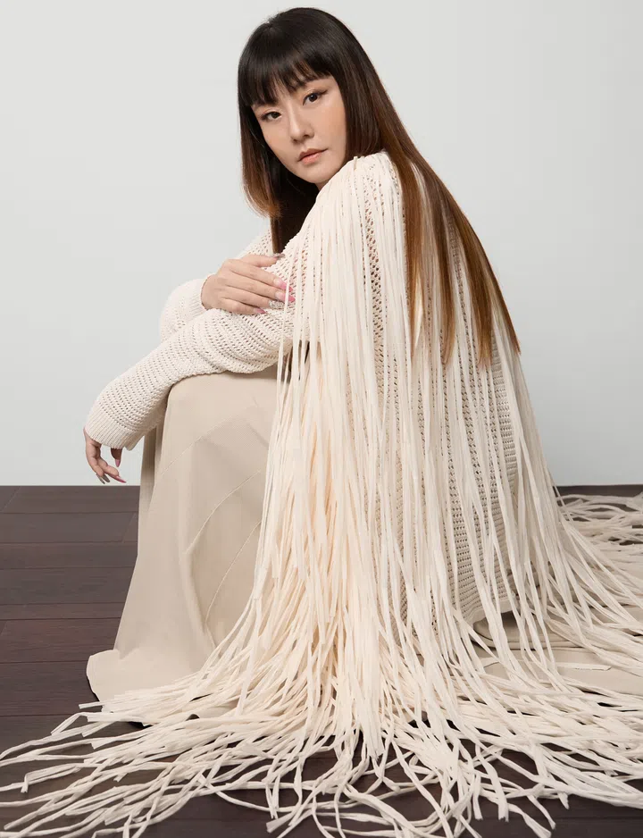 Yumiko Kayahara 坐在木质地板上，身穿米白色长流苏针织外套与浅色长裙，神情平静而专注，背景简洁明亮，展现柔和而自信的气质。
