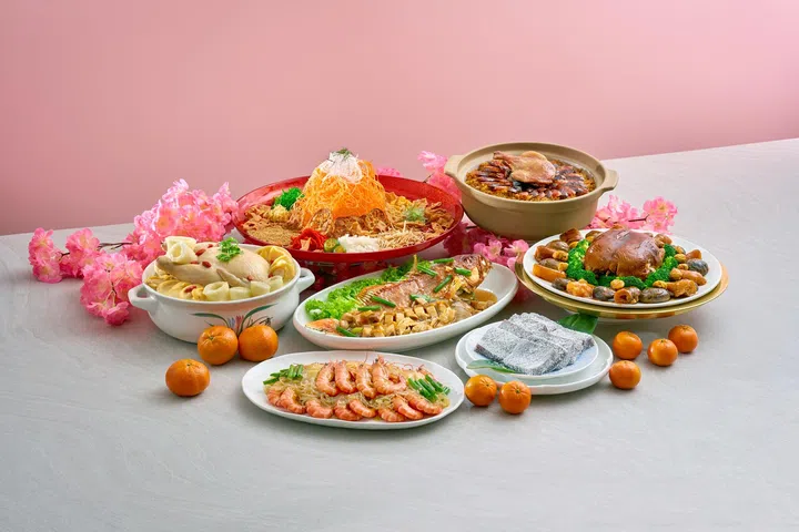 农历新年外带团圆年菜组合展示图，粉色背景与桌面上摆放多道中式佳肴，包括中央红盘盛装的捞起鱼生，周围搭配砂锅鸡汤、整条清蒸鱼、虾仁配面条、鲍鱼配西兰花的滋补菜肴，以及一锅丰盛盆菜，前方另有年糕甜点与多盘节庆菜式，桌面点缀橘子与粉色花卉装饰，整体呈现适合家庭聚餐的新加坡农历新年外卖团圆饭与节庆年菜氛围。