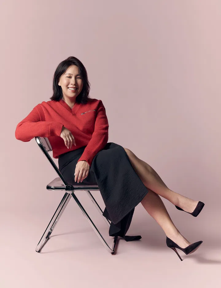 Charmaine Tan坐在金属椅上，身穿红色针织上衣与黑色长裙，脚踩黑色高跟鞋，微笑望向镜头。淡粉色背景营造出温暖柔和的氛围，展现她自信、亲切又坚定的气质。