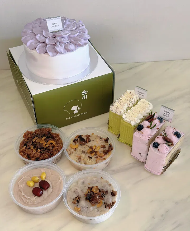 桌面上摆放 The Pine Garden 松园蛋糕与甜点组合，包括一款紫色花瓣装饰的整蛋糕，以及抹茶、莓果等切片蛋糕，旁边搭配多款传统甜品，呈现丰富多样的选择。2026母亲节蛋糕推荐：精致又好吃的限定款
