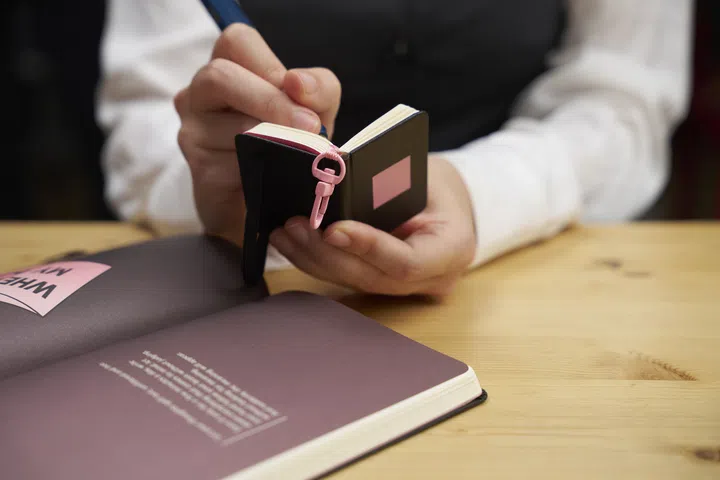 Moleskine 与 BLACKPINK 联名系列形象照，画面中一名使用者手持迷你笔记本书写，笔记本配有粉色 Notebook Charm 挂饰，桌面上摊开同系列大型笔记本，整体以温暖木质桌面与柔和色调呈现，展现结合 BLACKPINK 风格与 Moleskine 经典设计的限量联名文具，适合喜爱书写、手帐与明星联名的读者。
