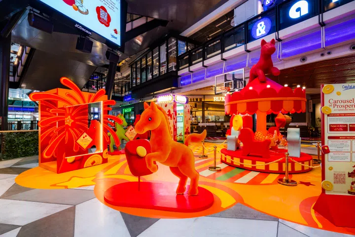 位于Funan商场中庭的“繁荣旋转木马”（Carousel of Prosperity）新春主题装置，以鲜艳的红色、橙色与金色为主色调，展现充满活力的马年节庆氛围。现场可见一匹立体骏马雕塑立于中央，象征马到成功与新年好运，旁边设有旋转木马造型装置，顶部同样饰有红色马匹雕像。周围搭配烟花造型灯饰、金元宝元素与节庆装饰，营造喜气洋洋的新春打卡空间。背景可见Funan商场现代化建筑结构、多层购物空间与商店灯光，整体呈现融合现代购物环境与传统农历新年元素的沉浸式节庆体验，吸引公众前来拍照留念，感受2026马年新春的欢乐气氛。