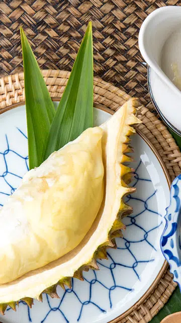 一盘榴莲与泰式甜品摆放在藤编桌布上，包括榴莲果肉、椰浆糯米与装有榴莲酱的白色小壶，旁边点缀着热带绿叶和白色鸡蛋花。