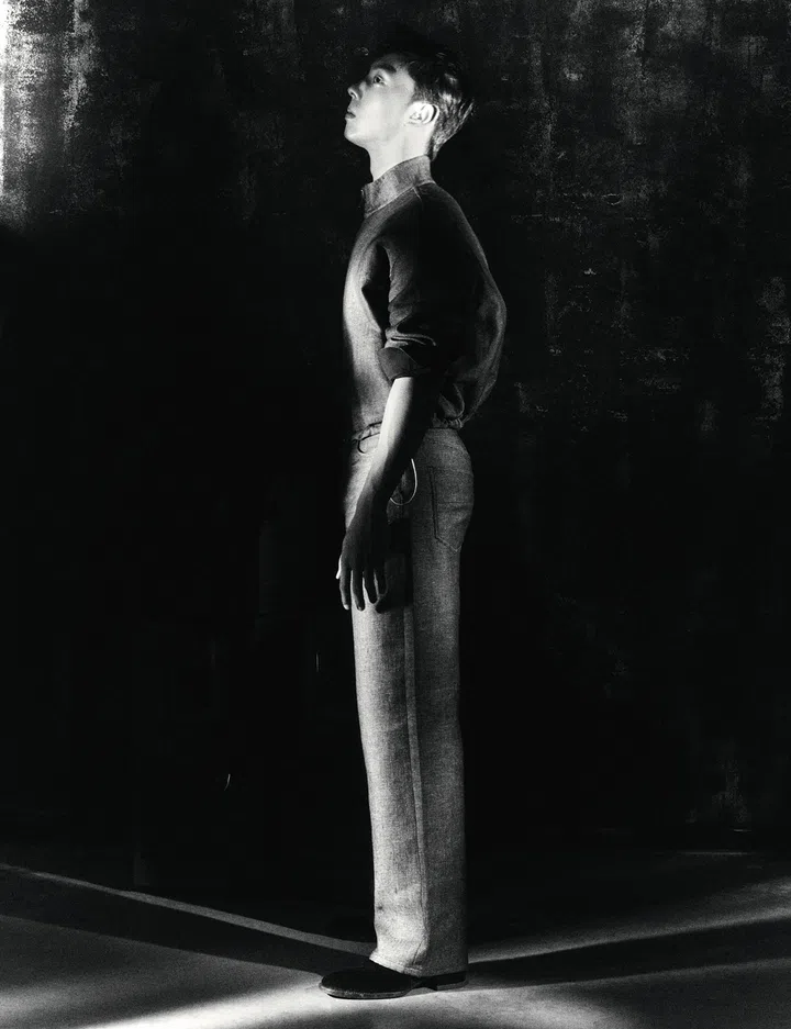 仲伟杰黑白时尚人像，侧身站立于暗色背景中，身穿简约服装，光影勾勒出修长身形与轮廓，展现冷静内敛的气质。