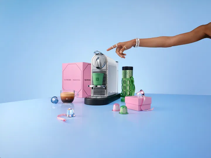 Nespresso与Swarovski携手推出2026节日限定联名系列，画面以淡蓝色为背景，中央展示点缀水晶装饰的银白色CitiZ Platinum咖啡机，机身前方摆放绿色咖啡杯，旁边搭配粉红色联名礼盒、绿色随行杯及饰有水晶的杯套。桌面散落粉红、绿色及水晶造型的咖啡胶囊装饰，左侧摆放一杯分层咖啡，右上方伸入一只佩戴闪亮水晶手链与戒指的手，展现Swarovski标志性的璀璨美学与Nespresso咖啡文化的结合。