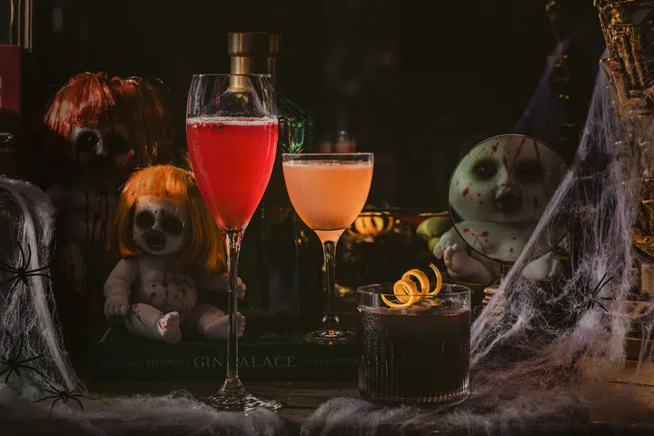 三杯万圣节主题鸡尾酒摆放在布满蜘蛛网与玩偶装饰的桌面上，营造神秘又华丽的节日氛围。新加坡万圣节去哪里玩推荐。