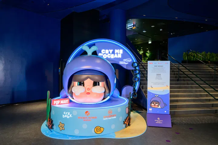 Singapore Oceanarium入口处展示“CRYBABY Cry Me an Ocean”主题装置，一个巨型CRYBABY角色戴着蓝色鲸鱼造型头套，双眼含泪，坐落于海洋主题圆形底座上。装置后方设有发光的“CRY ME an OCEAN”标志，并展示POP MART、CRYBABY、Resorts World Sentosa及Singapore Oceanarium标识。右侧设有介绍鲸鱼的主题立牌，整体以蓝色灯光和海洋元素营造沉浸式氛围，成为活动的热门拍照打卡点。