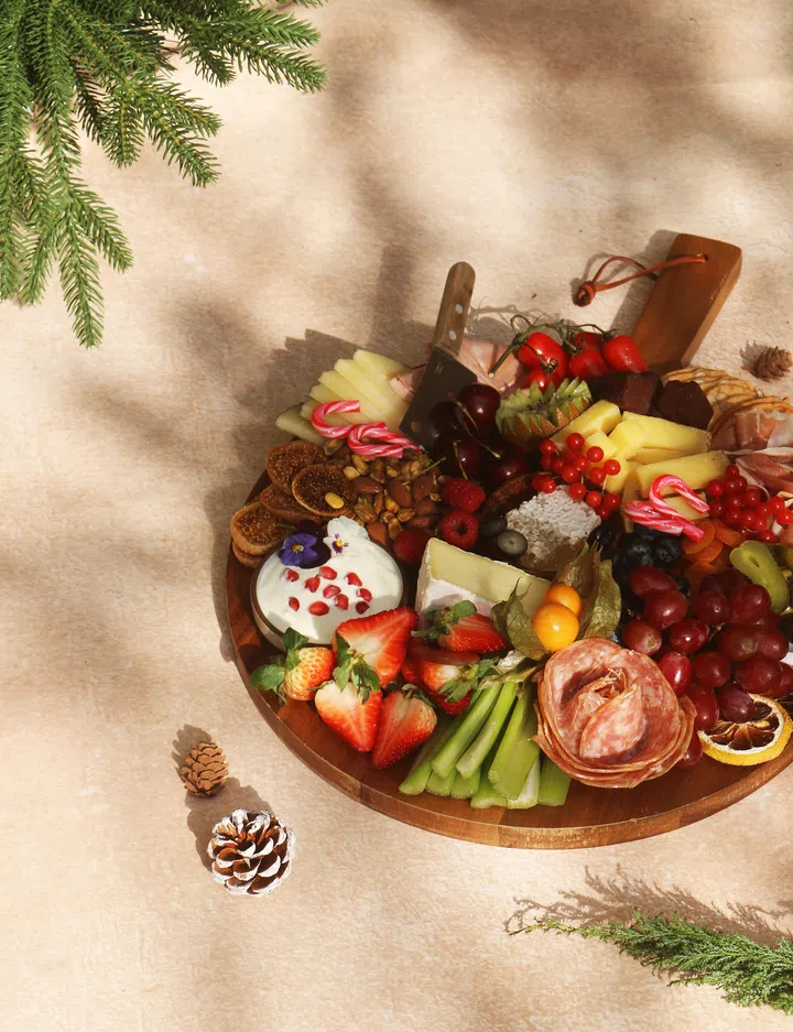 木盘上摆满圣诞主题的水果、奶酪、冷切肉、坚果、甜点和装饰小糖果，周围点缀松果与圣诞绿叶，整体呈现温暖又充满节庆气氛的圣诞拼盘画面。圣诞大餐吃什么？新加坡9家必订自助餐全攻略