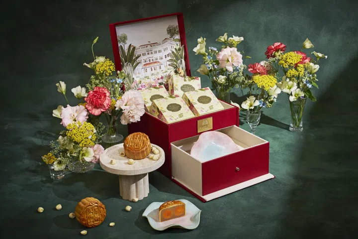 Raffles Hotel Singapore 2026中秋Premium Baked Mooncake Box礼盒场景图。酒红色双层礼盒打开后，上层摆放四盒独立包装月饼，盒盖内侧印有Raffles Hotel Singapore建筑插画；下层抽屉则放置一只粉蓝色陶瓷盘。前景陈列完整烘焙月饼及切开的双黄月饼，可清楚看到白莲蓉与两颗咸蛋黄内馅，四周以粉色、红色、黄色及白色鲜花装饰，整体呈现典雅精致的中秋送礼氛围。