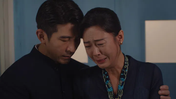 这是一张电视剧画面截图，呈现一段情绪浓烈的对手戏。画面中，一对男女紧靠在一起，神情沉重。女子神情崩溃，眼眶泛红，泪水顺着脸颊滑落，眉头紧锁，嘴角微微颤抖，明显处于极度悲伤与压抑的情绪之中。她身穿深色上衣，领口带有绿色刺绣细节，造型简约却带有生活气息。
男子则站在她身旁，侧脸微低，神情凝重且克制，一手轻轻搭在她的肩上，给予安慰与支持。他穿着深色立领上衣，整体造型沉稳内敛。两人的额头几乎贴近，形成一个亲密而充满情感张力的画面。
背景为室内空间，墙面为浅蓝色调，光线柔和但略带冷色，进一步烘托出画面的压抑氛围。整体构图紧凑，以人物面部表情为焦点，捕捉了情绪爆发的瞬间，让观者能够强烈感受到角色之间的关系与当下的情感重量。小娘惹之翡翠山张金河与二少奶奶李淑琴。刘子绚首夺视后：22年7次提名终圆梦，她把时间走成答案