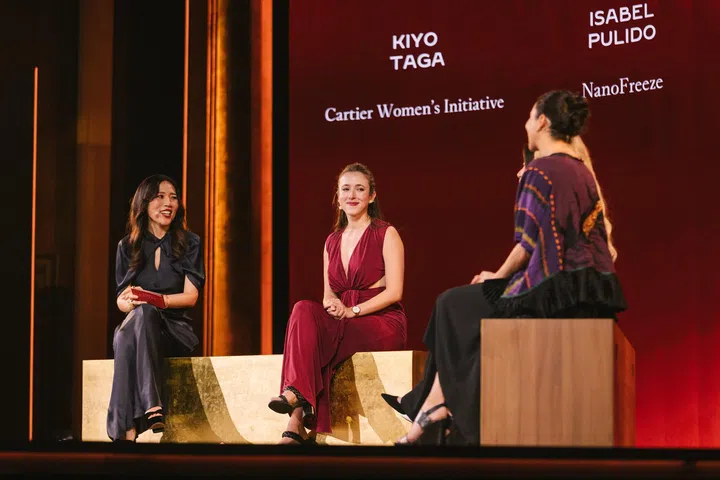 Cartier Women’s Initiative总监Kiyo Taga与两位女性创业者坐在舞台上进行对谈分享。三人分别坐在木质与金色座椅上，轻松交流创业经历、女性领导力及社会影响力等议题。背景大型屏幕显示讲者姓名及Cartier Women’s Initiative标志，现场论坛是20周年庆典的重要环节之一，邀请来自全球的创业者分享经验与交流想法。