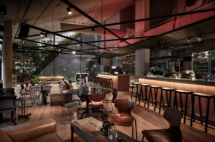 位于新加坡乌节路 Design Orchard 的 Third Floor 餐饮酒吧室内空间。场地采用现代工业风设计，设有宽敞的开放式吧台、木质高脚椅、圆形餐桌及舒适沙发座位。挑高天花板搭配黑色金属框架、轨道灯及大面积玻璃窗，室内以温暖的木质色调和柔和灯光营造轻松氛围，是举办2026万圣节与墨西哥亡灵节主题派对的场地。