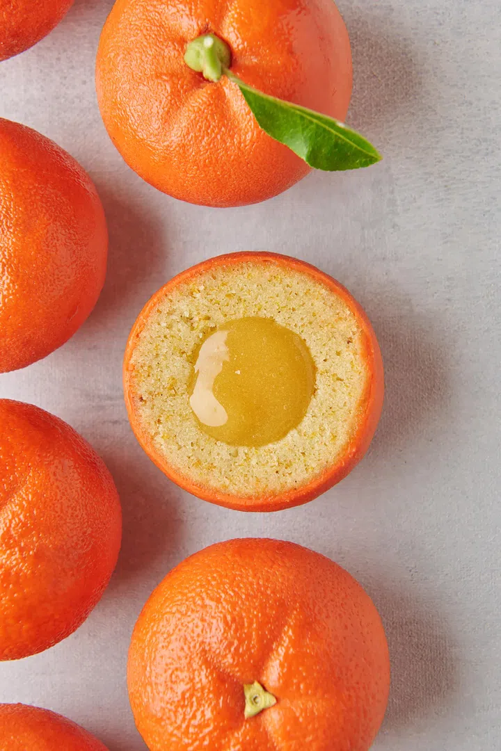 Cédric Grolet新加坡农历新年限定Mandarin柑橘甜点特写图。画面以多颗逼真柑橘造型甜点排列呈现，其中一颗被切开，露出细腻蛋糕层与金黄色果酱夹心，外观仿若真实橘子，带有绿色叶片点缀，整体色调明亮温暖，充满新年吉祥氛围。2026马年新年送礼推荐：拜年礼盒与新年礼物精选