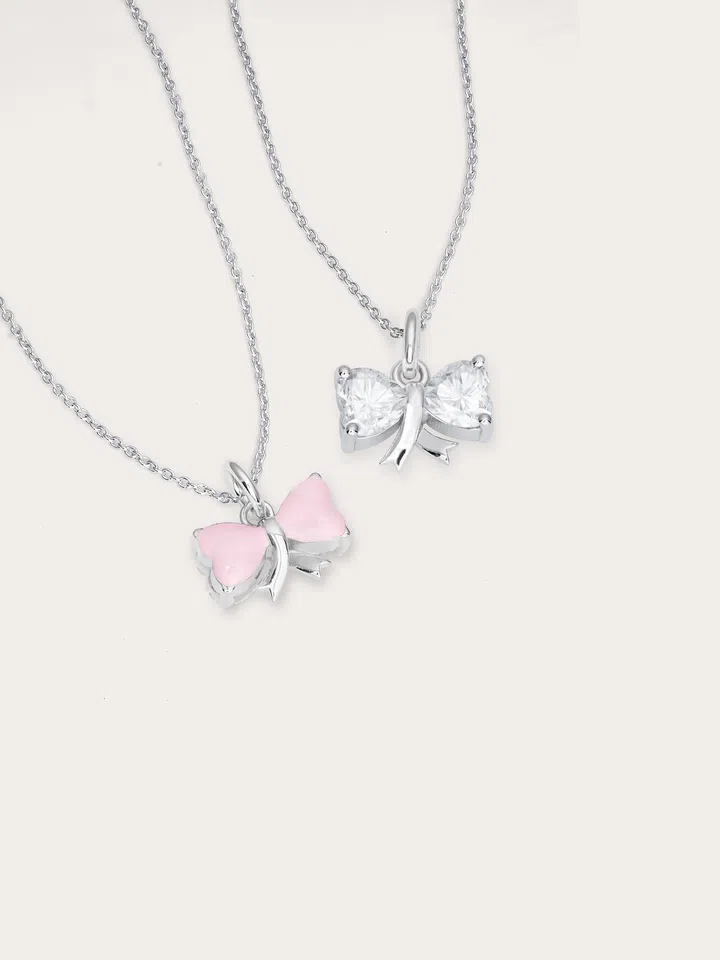 两条银色项链平铺在浅粉背景上，吊坠为蝴蝶结造型，其中一款饰以粉色珐琅，另一款镶嵌透明莫桑石，整体设计简约优雅，象征女性的温柔与坚韧。