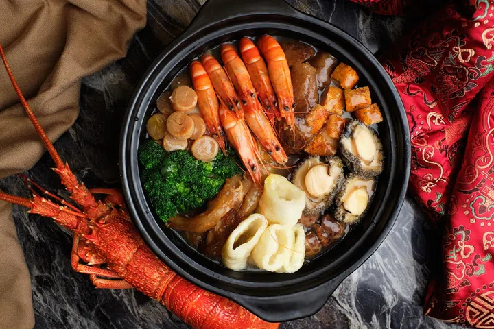 黑色砂锅中盛装海鲜盆菜，整齐摆放多只整虾、鲍鱼、干贝、花胶、鱼鳔与西兰花等配料，汤汁浓郁，食材分区排列，周围搭配红色新年布饰与龙虾装饰，营造热闹喜庆的农历新年用餐氛围，新加坡盆菜推荐2026 农历新年必吃餐厅与价格整理。