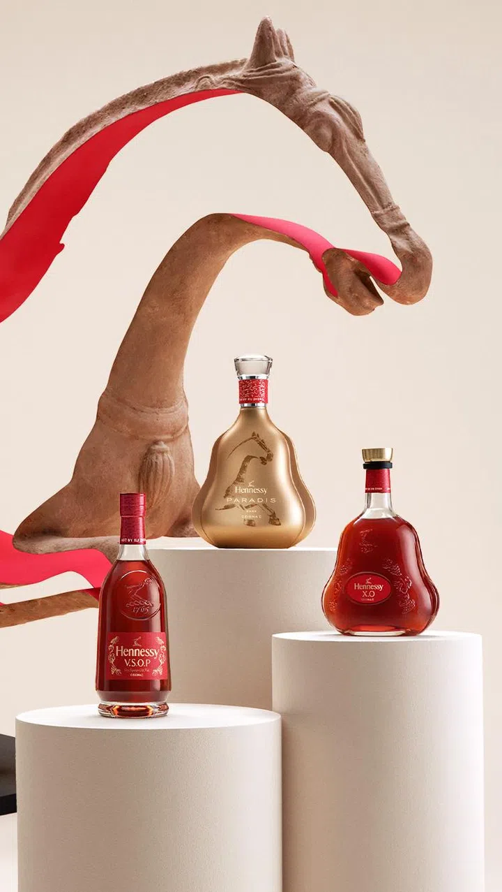 Hennessy 2026马年限量版干邑系列展示图。画面以简约艺术风格呈现三款经典酒款——Hennessy V.S.O.P、Hennessy X.O与Hennessy Paradis，分别陈列于白色圆柱展台上。酒瓶设计融入喜庆红色与金色元素，背景可见带有马形轮廓的装置艺术，象征马年活力与力量，整体构图现代优雅，适合作为高端新年送礼之选。2026马年新年送礼推荐：拜年礼盒与新年礼物精选