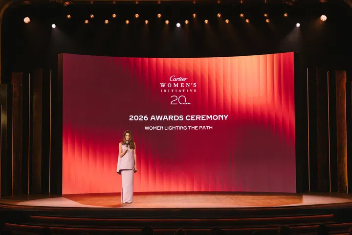 Cartier Women’s Initiative 20周年颁奖礼主舞台以红橘色渐层LED屏幕作为背景，画面中央展示“Cartier Women’s Initiative 20 Years”、“2026 Awards Ceremony”及“Women Lighting the Path”等字样。一名主持人站在舞台中央手持麦克风主持活动，聚光灯照亮整个舞台，象征庆祝20周年的重要里程碑。