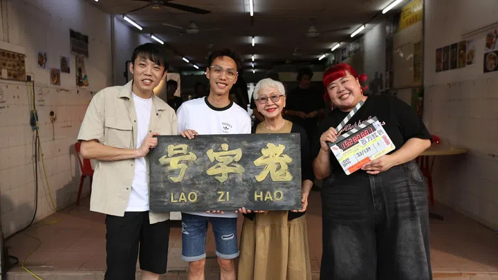 《百万红包 A Good Fortune》拍摄现场幕后合照，导演 Jason Lee 与演员王伟良、Usha Seamkhum 及 Xixi Lim 站在咖啡店内合影，众人面带笑容，一起举着写有“老字号 Lao Zi Hao”的招牌与电影打板，背景可见拍摄器材与剧组人员，呈现电影拍摄阶段的重要时刻与团队完成一场戏后的合照氛围。